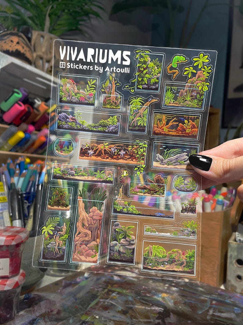 May include: Clear sticker sheet featuring colorful vivarium designs. The sheet includes various rectangular and circular stickers with detailed illustrations of terrarium landscapes, plants, and small animals. The text "Vivariums" and "Stickers by Artoulli" are printed on the sheet.