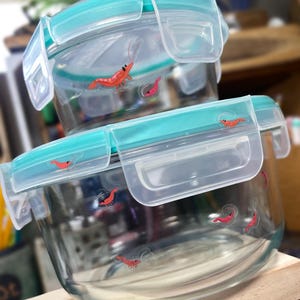 May include: Two round clear glass food storage containers stacked vertically. Each container features a teal plastic lid with four locking tabs and a pattern of small red shrimp illustrations printed on the glass and the lid surfaces.