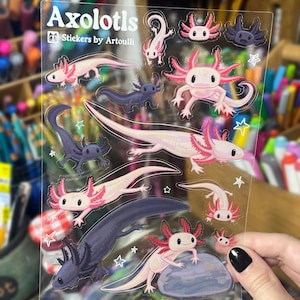 以下が含まれることがあります： ピンク、白、濃い青色のアホロートルデザインが施された透明なステッカーシート。「Axolotls」と「Stickers by Artoulli」の文字入り。様々なポーズとサイズのウーパールーパーと星のアクセント。