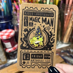 May include: A novelty pin featuring a green frog wearing a wizard hat with stars. The pin is on a brown card with the text "PAOLO THE PRESTIDIGATOR MAGIC MAN". The card also includes the text "FRIDAY 9PM ARTOULLI LLC".