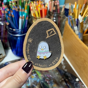 Puede incluir: Un pin de esmalte blanco con un diseño de un pájaro pequeño de color morado claro y verde con patas amarillas. El pin está en una tarjeta marrón con el texto "enamel pin by artoulli on ETSY".