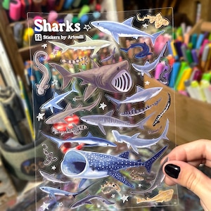 May include: Clear sticker sheet featuring various shark illustrations in shades of blue, gray, and brown. The sticker sheet includes the text "Sharks" and "Stickers by Artoulli".