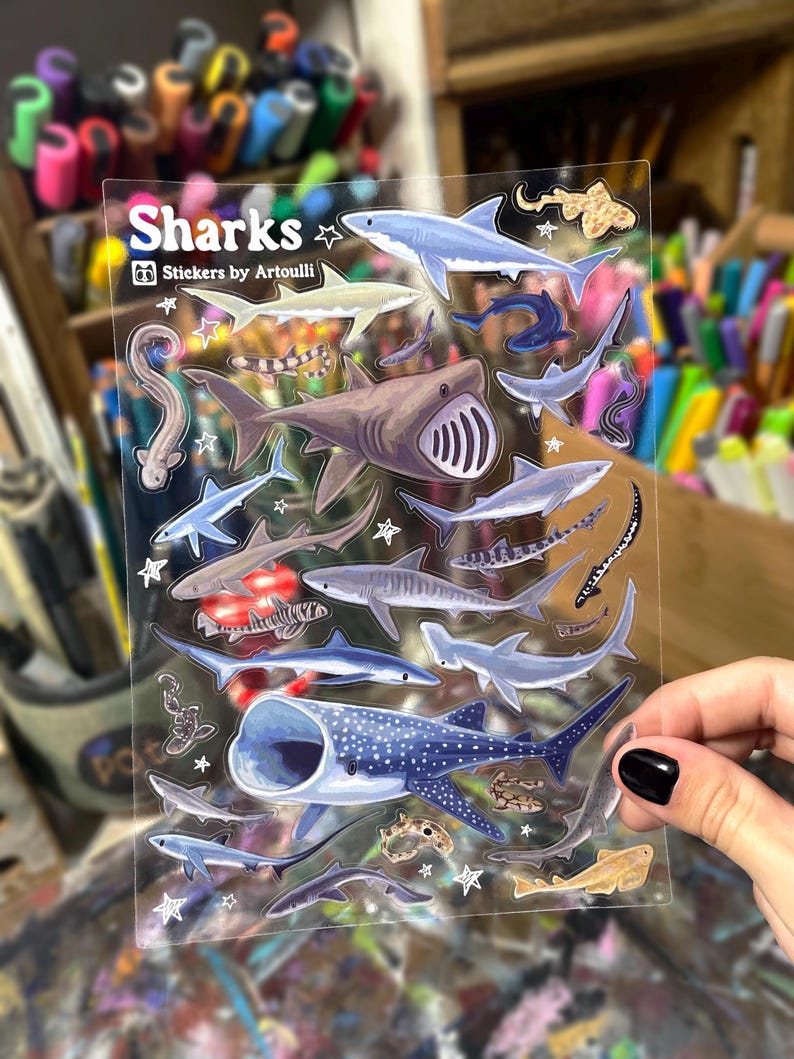 May include: Clear sticker sheet featuring various shark illustrations in shades of blue, gray, and brown. The sticker sheet includes the text "Sharks" and "Stickers by Artoulli".