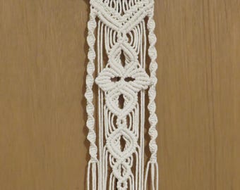May include: Off-white macrame wall hanging with a geometric design. The piece is suspended from a piece of driftwood and features intricate knotting patterns, including diamond and spiral details, with long fringe.