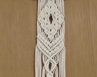 May include: Cream-colored macrame wall hanging. The design features a geometric pattern with diamond shapes and a fringe of vertical cords. The macrame is attached to a natural wooden dowel, creating a bohemian aesthetic.