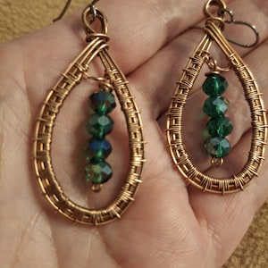 May include: A pair of teardrop-shaped earrings crafted from coiled copper wire. Each earring features a central vertical strand of four faceted, dark green crystal beads suspended within the wire frame, attached to a simple metal hook ear wire.