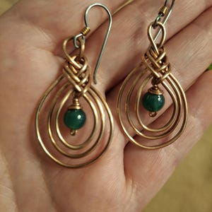 May include: A pair of dangle earrings featuring a teardrop-shaped wire frame made of copper-toned metal. Each earring has a central green bead suspended within the wire loops and is attached to a silver-toned metal ear wire hook.