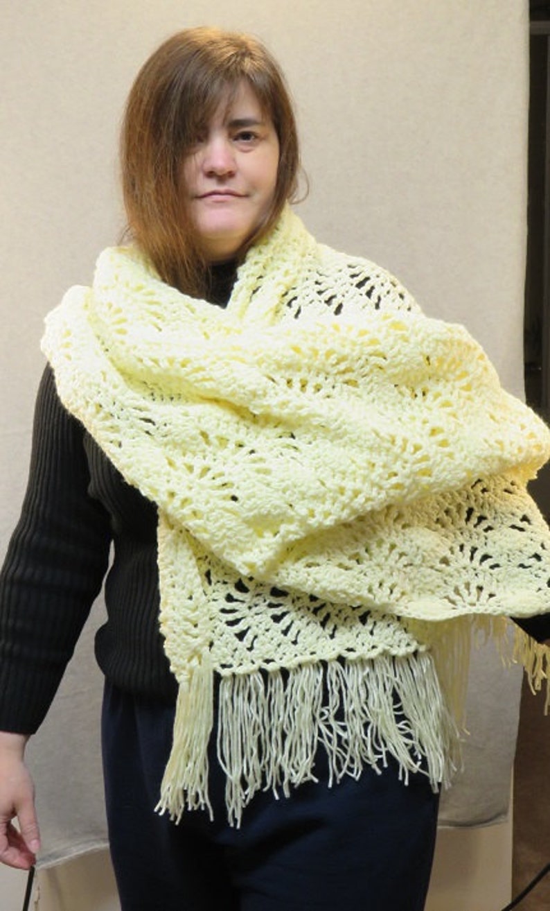 May include: A woman wearing a pale yellow crocheted shawl with fringe. The shawl is draped around her shoulders and neck.