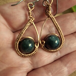 May include: A pair of wire-wrapped dangle earrings featuring a teardrop-shaped copper frame. Each earring contains a single, smooth, dark green spherical bead secured within the center of the coiled copper wire structure, attached to a silver-toned hook.