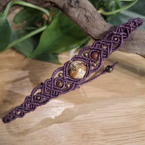 May include: A macrame bracelet crafted from dark purple cord featuring an intricate knotted pattern. The design incorporates a central round beige and brown patterned stone bead, accented by smaller metallic bronze beads and dark brown stone beads.