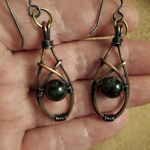 May include: A pair of dangle earrings featuring a dark green spherical gemstone encased in a teardrop-shaped copper wire frame. The wire is wrapped with intricate coils and attached to a dark metal hook for pierced ears, showcasing a rustic finish.