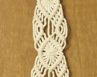 May include: Cream-colored macrame wall hanging with a wooden ring at the top. The design features geometric patterns and a fringe at the bottom. This handmade piece adds a bohemian touch to any space.