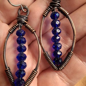 May include: A pair of dangle earrings featuring a marquise-shaped frame made of dark, oxidized copper wire. Each earring contains a vertical row of six faceted, deep blue crystal beads suspended within the center of the wire frame.