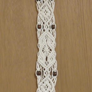 May include: Cream-colored macrame wall hanging with a geometric design. The woven piece features dark brown wooden beads and a natural wooden rod at the top for hanging. The macrame has a symmetrical pattern.