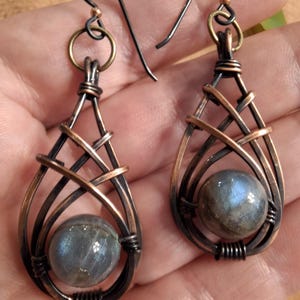 May include: A pair of wire-wrapped earrings featuring teardrop-shaped frames made from oxidized copper wire. Each earring contains a round, iridescent labradorite gemstone at the base, secured by intricate wire coils and suspended from dark metal hooks.