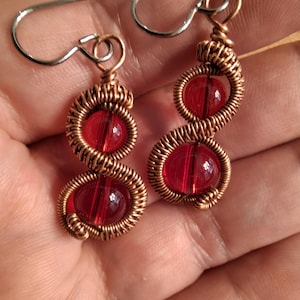 May include: A pair of dangle earrings featuring two round, translucent red glass beads wrapped in copper wire. The wire forms a figure-eight shape around the beads, and each earring is attached to a silver-toned metal hook for ear piercing.