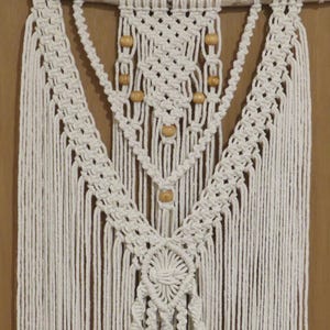 May include: Cream-colored macrame wall hanging. The design features intricate knotting patterns, wooden beads, and a fringe. The macrame is suspended from a natural wooden branch, creating a bohemian aesthetic. A decorative piece for home decor.