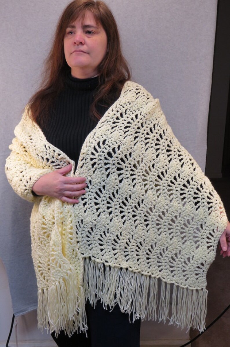 May include: A pale yellow crocheted shawl with a delicate openwork pattern and fringe. The shawl is draped over a person's shoulders.