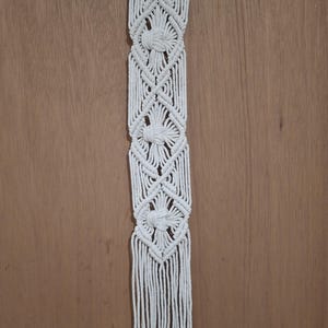 May include: A white macrame wall hanging featuring intricate knotted diamond patterns and a central vertical row of three decorative knots. The piece is suspended from a natural driftwood branch with a white cord loop for hanging at the top.