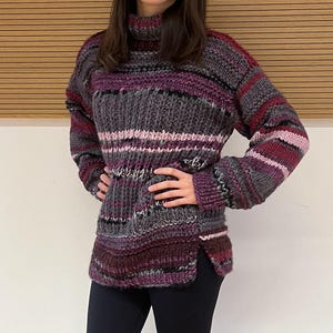 Könnte beinhalten: Ein gestreifter Rollkragenpullover in den Farben Lila, Grau und Rosa. Der Pullover ist aus dickem Garn gestrickt und hat eine lockere Passform.