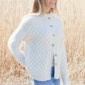 Könnte beinhalten: Ein weißer Strickcardigan mit Knopfleiste und einem Zopfmuster. Der Cardigan wird an einem Model vor einem verschwommenen Hintergrund aus hohem Gras gezeigt.
