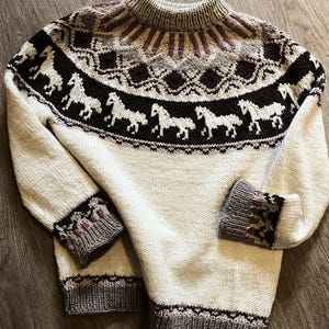 Könnte beinhalten: Cremefarbener Strickpullover mit hohem Kragen und langen Ärmeln. Das Design zeigt eine Reihe weißer Pferde auf dunklem braunen Hintergrund, mit geometrischen Mustern in Braun-, Grau- und Burgundertönen um Hals und Bündchen. Der Pullover ist aus Strickmaterial.