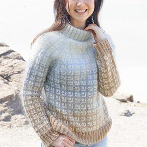 Könnte beinhalten: Ein hellblauer und brauner Pullover mit einem hohen Kragen. Der Pullover ist aus weichem, flauschigem Garn gefertigt. Das Model trägt den Pullover mit einer blauen Jeans. Der Text "DROPS DESIGN" und "www.garnstudio.com" ist am unteren Rand des Bildes sichtbar.