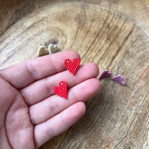 Könnte beinhalten: Zwei kleine rote herzförmige Ohrringe aus Perlen. Die Ohrringe werden in einer Hand vor einem Holzgrund gehalten.