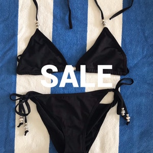 May include: Black bikini set with silver embellishments. The top is a triangle style and the bottom is a tie-side style. The word "SALE" is written in white letters across the image.