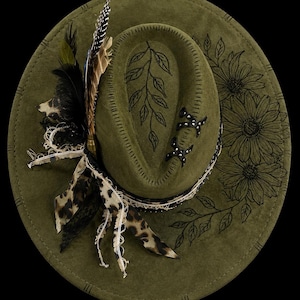 Puede incluir: Un sombrero fedora de fieltro verde oliva con un diseño de hojas dibujado a mano en la copa e ilustraciones florales en el ala. El sombrero está adornado con una cinta de estampado de leopardo, encaje deshilachado, plumas y dos detalles en forma de X.