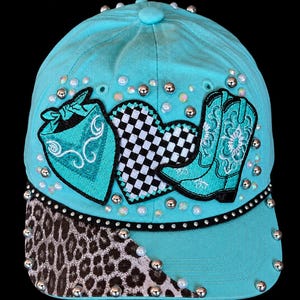 May include: A bright turquoise baseball cap featuring a black and white checkered heart, a turquoise bandana patch, and a turquoise cowboy boot patch. The brim has a partial leopard print pattern, and the entire cap is adorned with silver studs.
