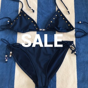 May include: A navy blue bikini with gold accents. The top is a triangle style with adjustable straps and the bottom is a tie-side style.