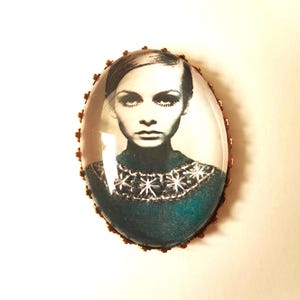 May include: Oval-shaped glass brooch with a gold-toned metal frame. The brooch features a black and white image of a woman with dark hair and a teal sweater with a white snowflake pattern.
