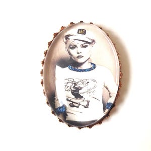 Op de afbeelding: Ovale broche met een zwart-wit afbeelding van een persoon met een matrozenpet en een wit shirt met een cartoonafbeelding. De broche heeft een bronskleurige, getextureerde rand en een transparante beschermhoes.
