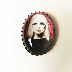 Op de afbeelding: Een ovale broche in vintage stijl met een zwart-wit afbeelding van een vrouw met blond haar. De afbeelding is geplaatst op een roze stoffen achtergrond en omlijst met een goudkleurige metalen rand.