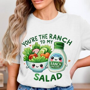 May include: White t-shirt featuring a graphic of a salad bowl and a bottle of ranch dressing. The text on the shirt reads "You're the ranch to my salad."
