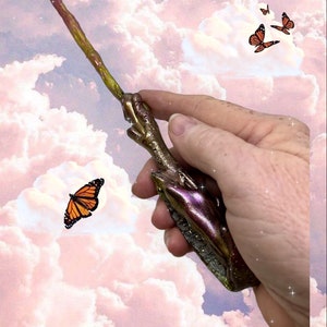 May include: A handcrafted wand with a metallic finish, featuring shades of gold, purple, and green. The wand is held against a backdrop of a cloudy sky with orange and black butterflies. This magical item is perfect for cosplay or fantasy enthusiasts.