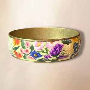 May include: A wide brass bangle with a floral design. The bangle is painted with a variety of flowers in shades of pink, purple, blue, yellow, and orange on a light green background.