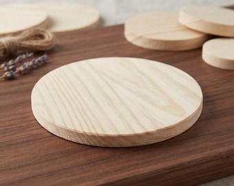 Puede incluir: Un conjunto de posavasos circulares de madera de color claro con un patrón de veta natural visible. Los posavasos están dispuestos sobre una tabla de madera marrón oscuro, con algunas piezas apiladas y otras planas, mostrando su superficie lisa.