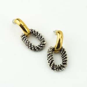 Puede incluir: Un par de pendientes con un lazo superior dorado y un colgante ovalado plateado. El colgante ovalado tiene un diseño de cuerda trenzada. Los pendientes se muestran sobre un fondo blanco.