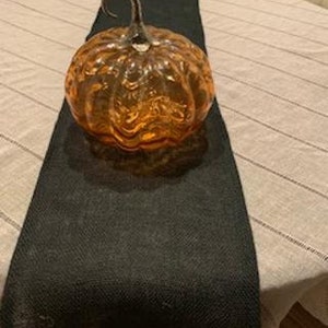 May include: A glass pumpkin with a textured surface and a silver stem. The pumpkin is orange and is sitting on a black table runner.