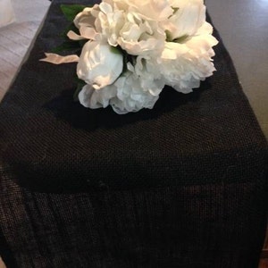 May include: A black burlap table runner with a bouquet of white peonies on top.