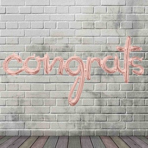 May include: Rose gold foil balloons spelling out the word "congrats" against a white brick wall background. A wooden floor is in the foreground.