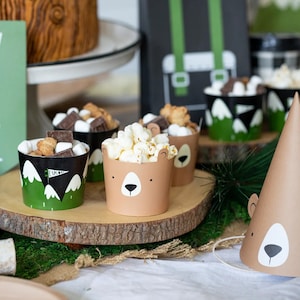 May include: A woodland-themed party table setting with brown and green paper cups filled with popcorn and snacks. There is a brown paper party hat with a bear face on it.