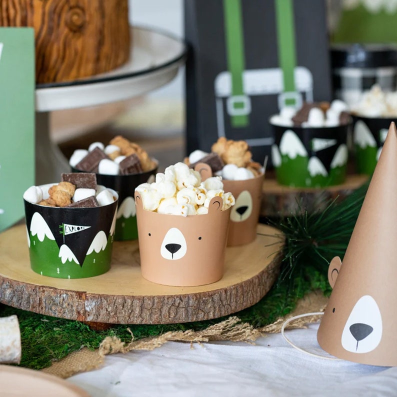 May include: A woodland-themed party table setting with brown and green paper cups filled with popcorn and snacks. There is a brown paper party hat with a bear face on it.