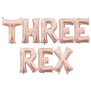 May include: Rose gold foil letter balloons spelling out "THREE REX".