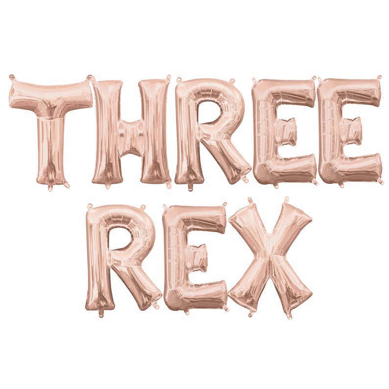May include: Rose gold foil letter balloons spelling out "THREE REX".