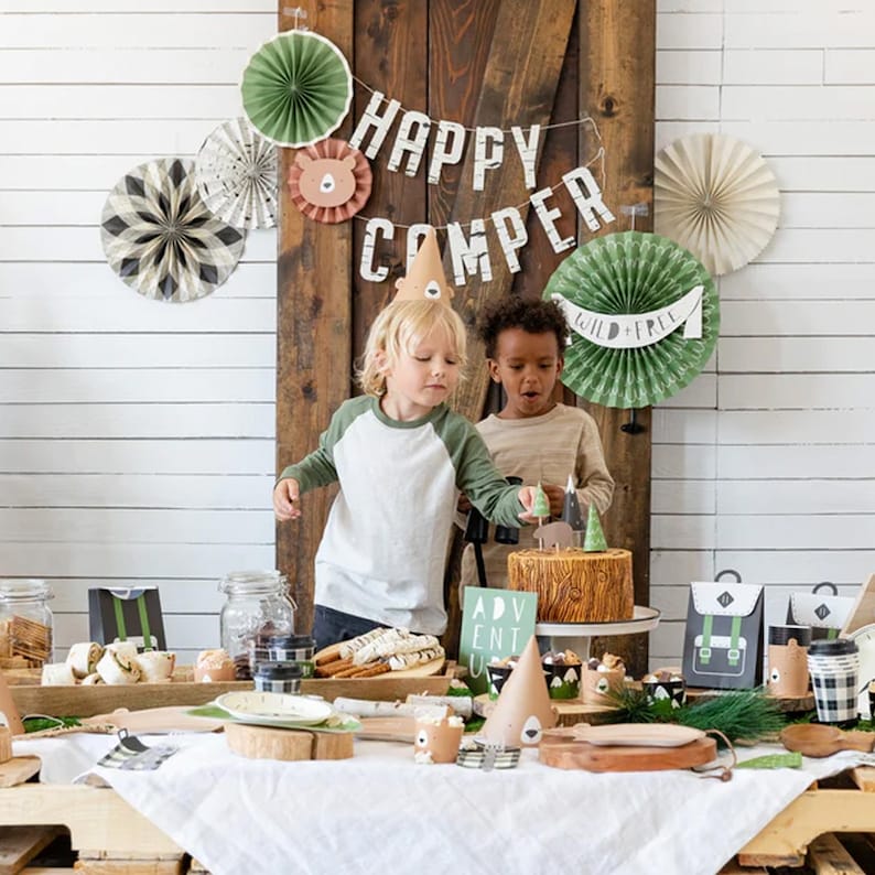 May include: A woodland-themed birthday party with a cake decorated like a tree stump, a banner that says "Happy Camper", and paper fans in green and brown. Two children are wearing brown bear party hats and are looking at the cake.