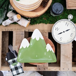May include: Green paper plate shaped like a mountain with white snow peaks and small green trees. The plate is on a wooden surface with a black and white checked napkin that says "Adventure".