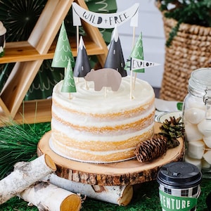 May include: A white cake with vanilla frosting decorated with a brown bear, green trees, and a banner that reads "Wild + Free". The cake is on a wooden cake stand and surrounded by green moss.